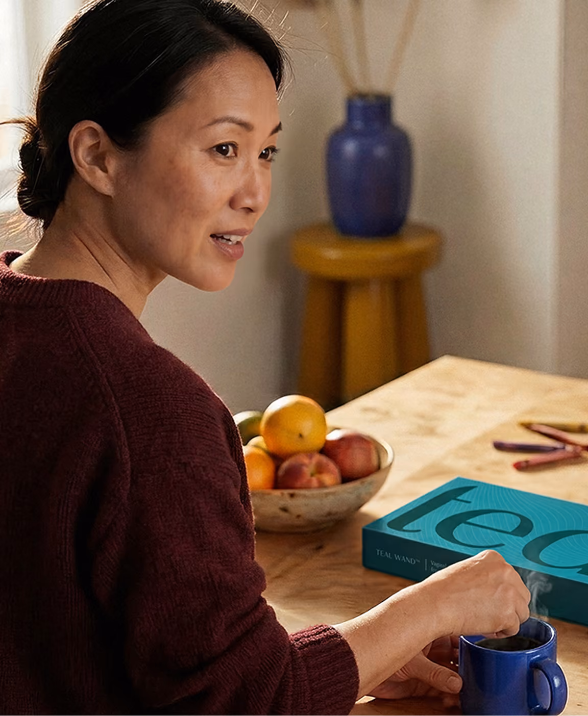 Woman in a maroon sweater sits at a wooden table with a bowl of fruit and a Teal box, holding a blue mug.