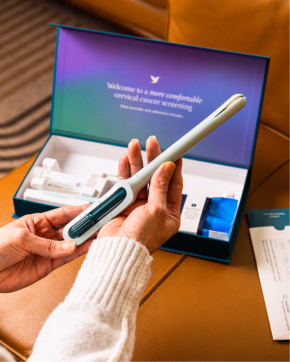 A close-up view of a person's hands holding the Teal Wand self-collection device over an opened kit box placed on a tan leather couch.