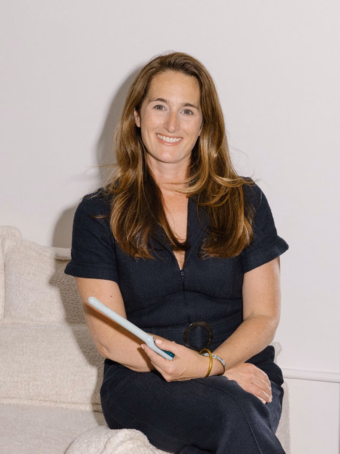 A professional portrait of Kara Egan, CEO and Co-founder of Teal Health, sitting on a white couch and smiling at the camera while holding the Teal Wand at-home cervical cancer screening device.