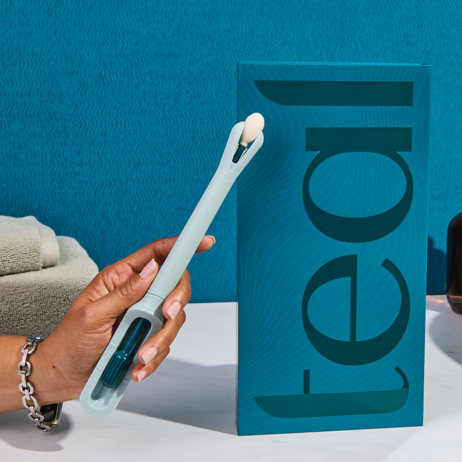 A hand holds up the light-teal colored Teal Wand self-collection device next to its vertical product packaging on a white marble bathroom vanity, showcasing its modern, user-friendly design.