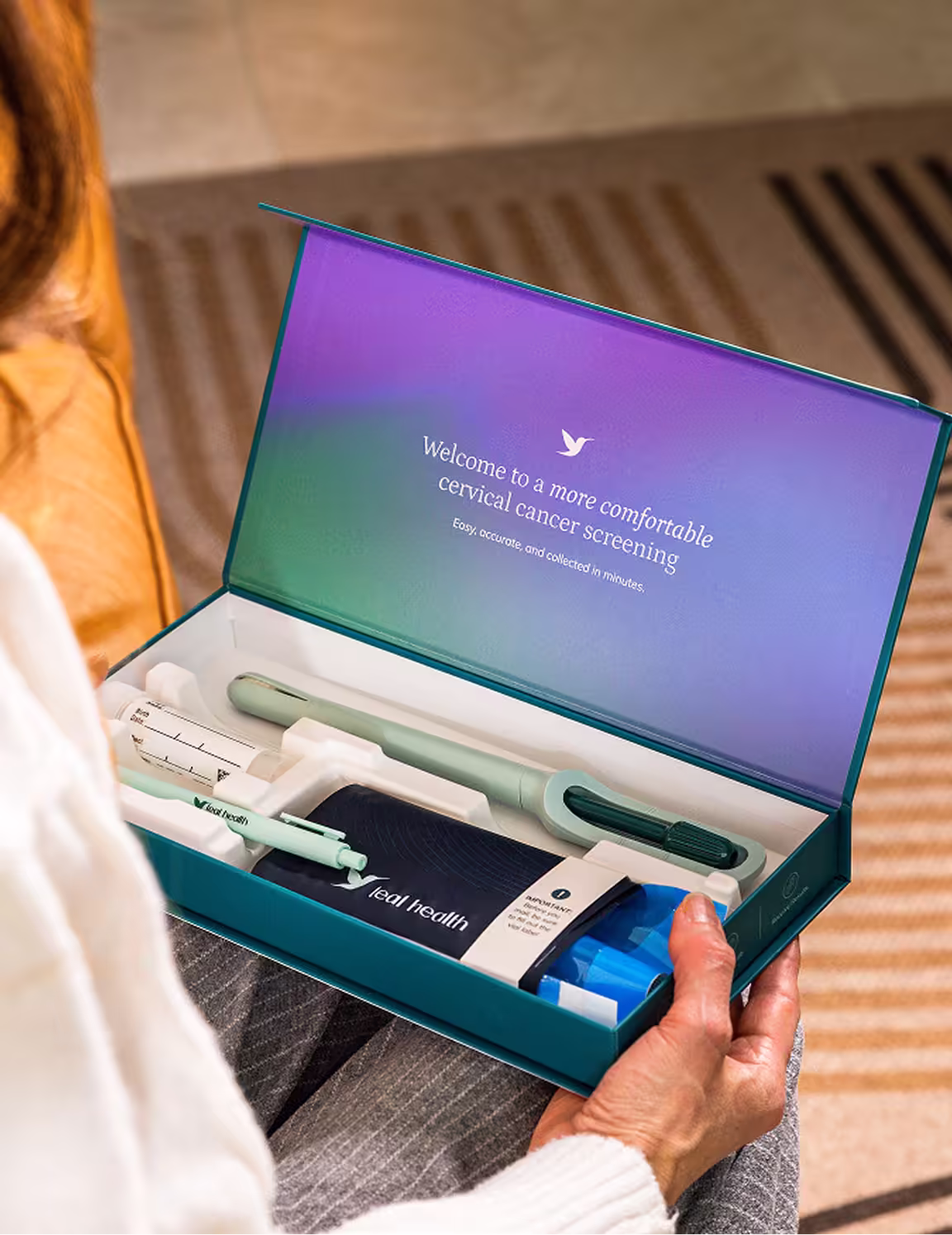 An over-the-shoulder view of a woman unboxing her Teal Health at-home cervical cancer screening kit. The opened box displays the Teal Wand device, a collection vial, a return mailer envelope, and an inside lid reading "Welcome to a more comfortable cervical cancer screening."