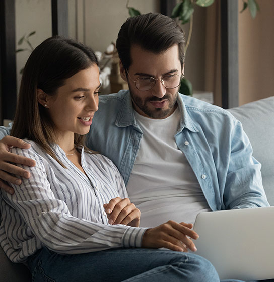 A woman and a man sit close on a couch, looking together at an open laptop.