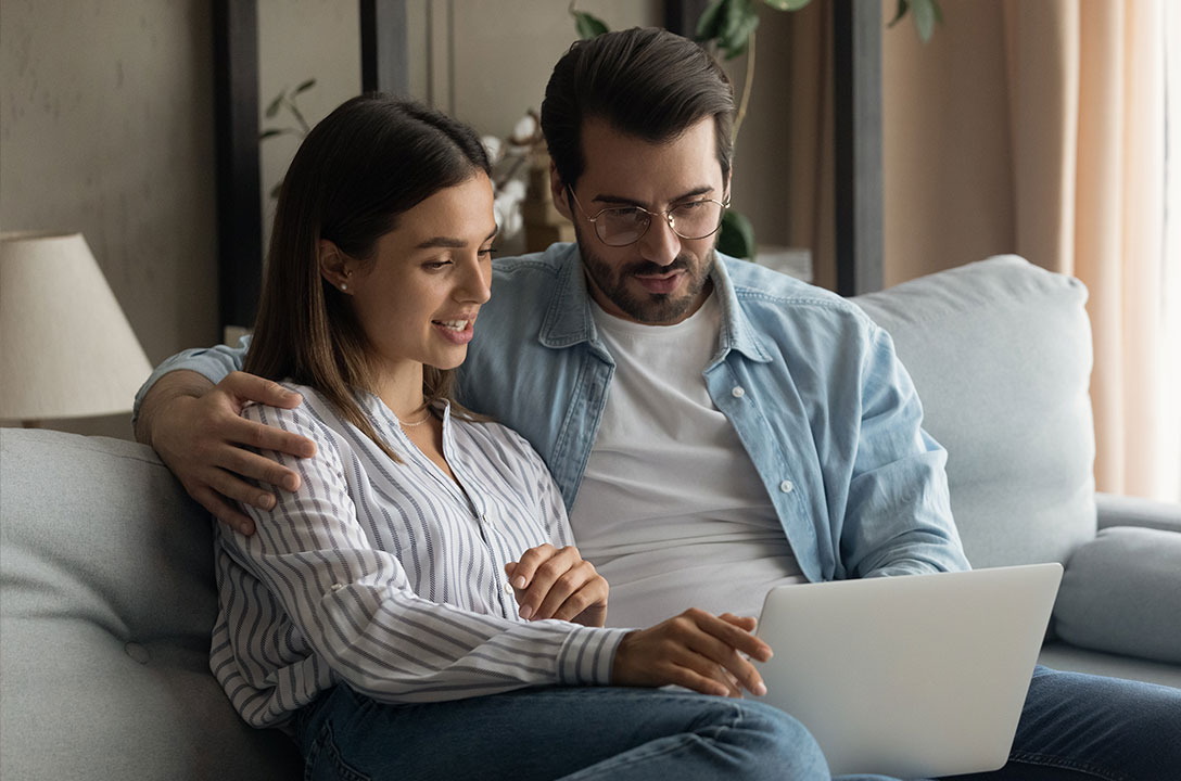 A woman and a man sit close on a couch, looking together at an open laptop.