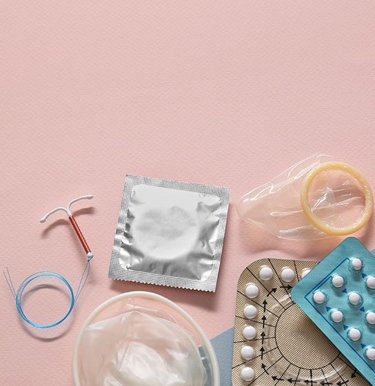 Flat lay of birth control methods on pink: IUD, condoms, pill packs, and a diaphragm.