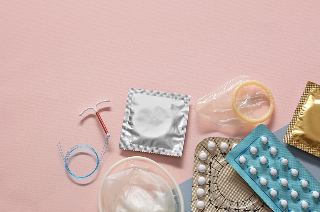 Flat lay of birth control methods on pink: IUD, condoms, pill packs, and a diaphragm.