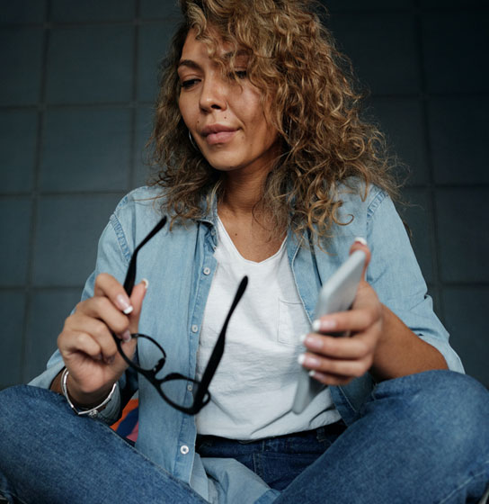 Curly-haired woman sits cross-legged holding eyeglasses and a phone in front of an open laptop.