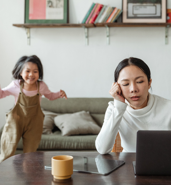 Tired woman rests her head on her hand at a laptop as two children run and play behind her.