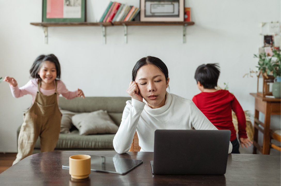 Tired woman rests her head on her hand at a laptop as two children run and play behind her.