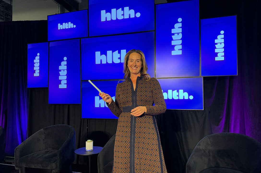 Smiling woman in a patterned dress holds a thin white wand on a stage lit by screens reading 'hlth.'