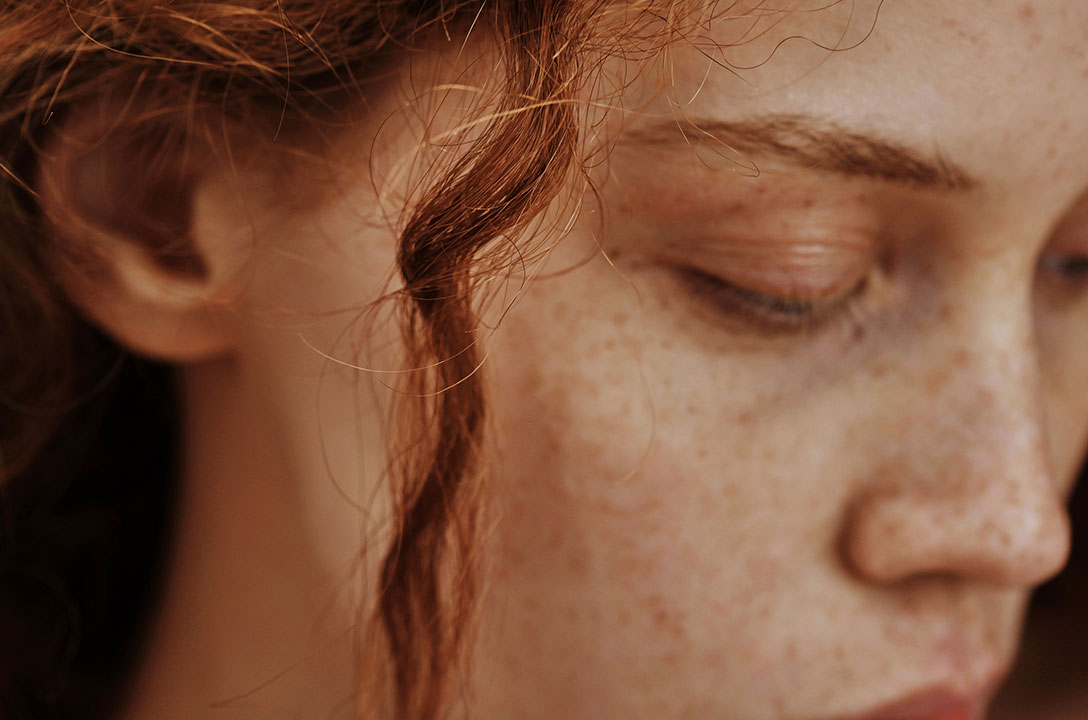 Close-up of a freckled face with red hair and eyes cast downward.