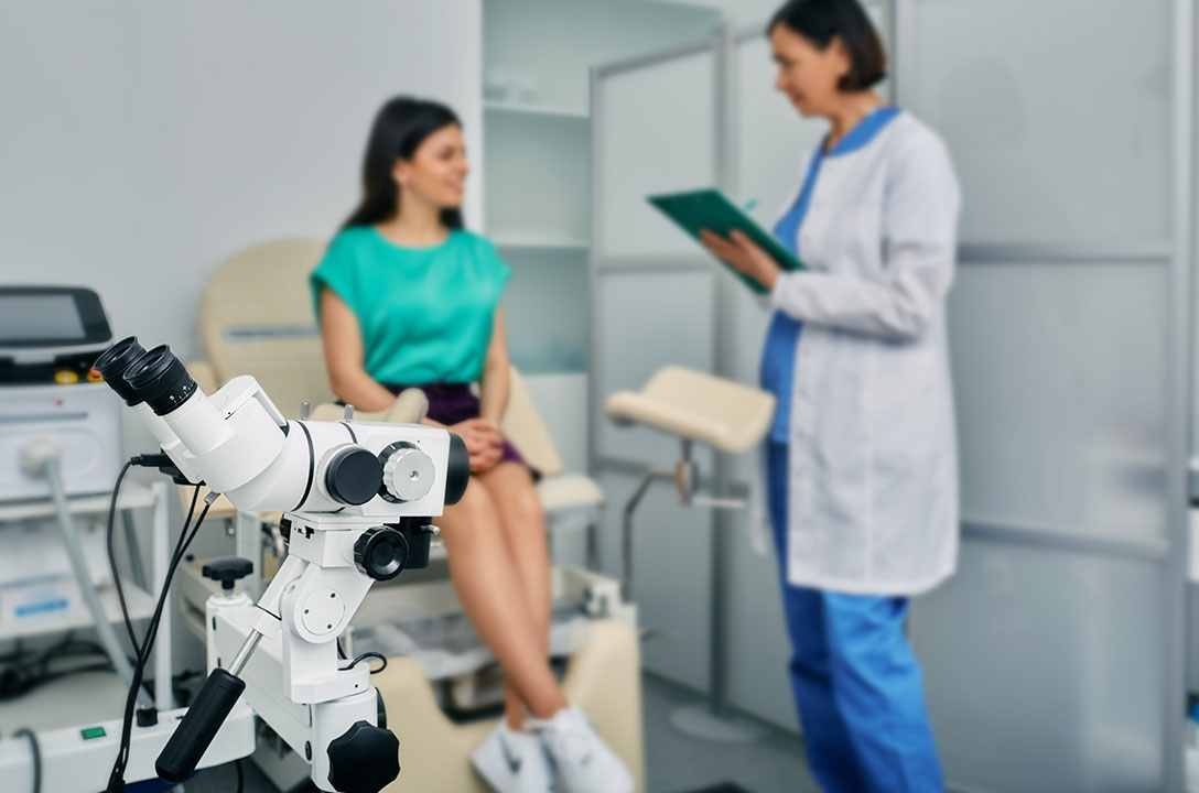 Clinic exam room with a colposcope in front, a patient in teal seated, and a clinician holding a clipboard.