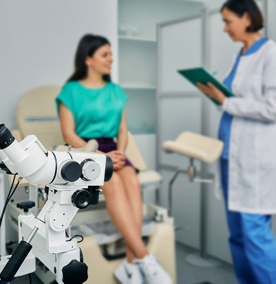 Clinic exam room with a colposcope in front, a patient in teal seated, and a clinician holding a clipboard.