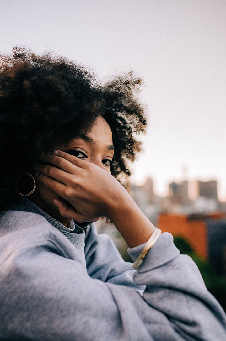 Woman with curly hair and gold hoop earrings resting a hand near her face, blurred city skyline behind.