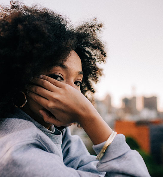 Woman with curly hair and gold hoop earrings resting a hand near her face, blurred city skyline behind.