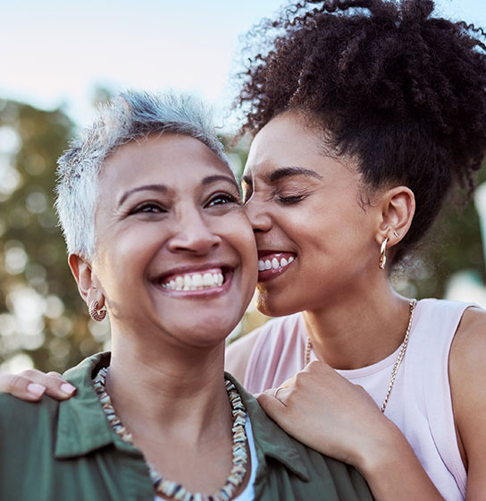 Two smiling women outdoors, one with short gray hair, the younger nuzzling her cheek.