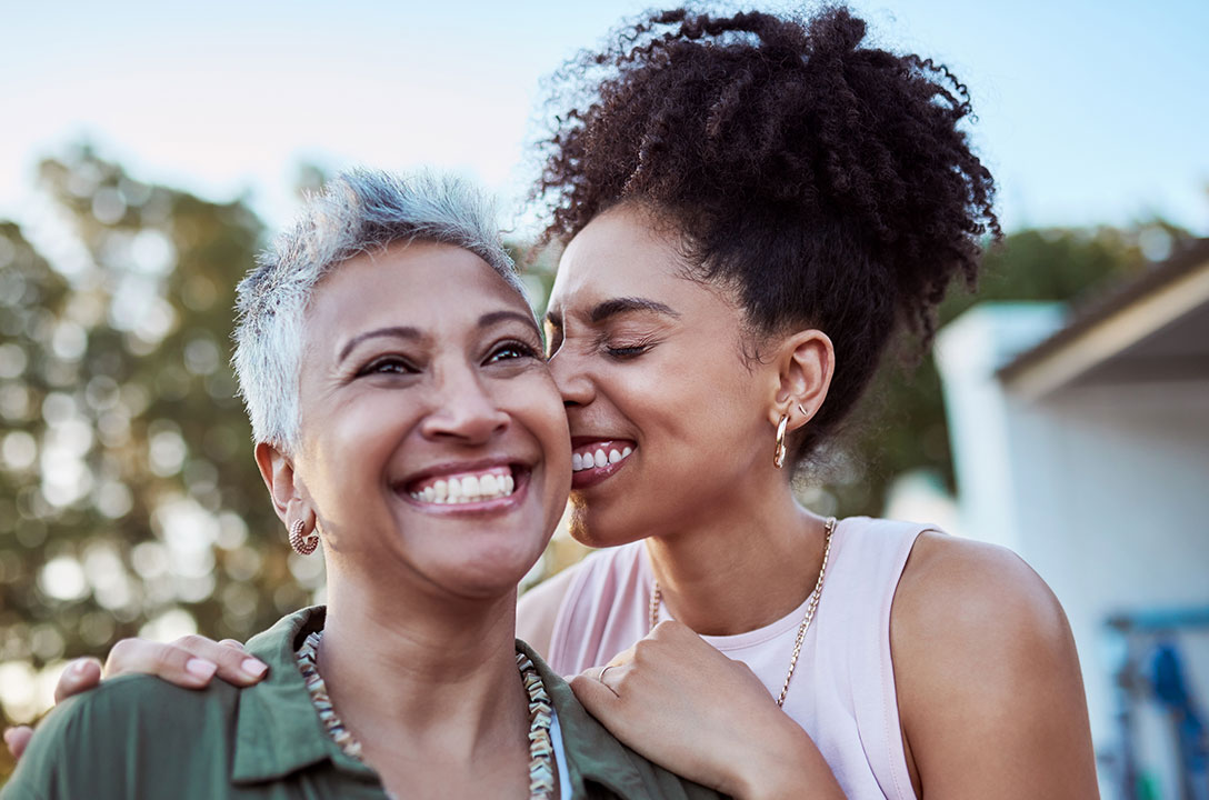 Two smiling women outdoors, one with short gray hair, the younger nuzzling her cheek.