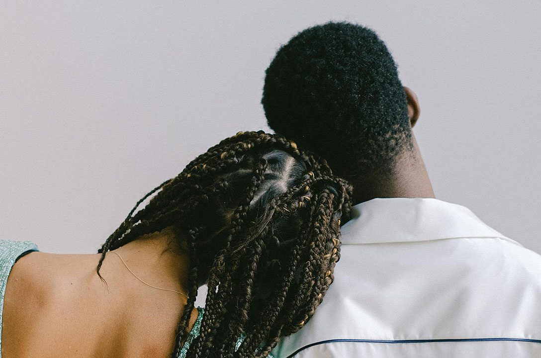 Back view of a woman with long braids resting her head on the shoulder of a man in a white shirt.