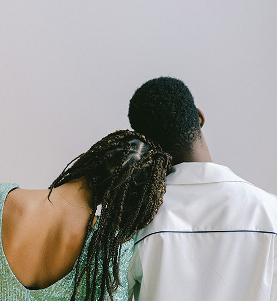 Back view of a woman with long braids resting her head on the shoulder of a man in a white shirt.