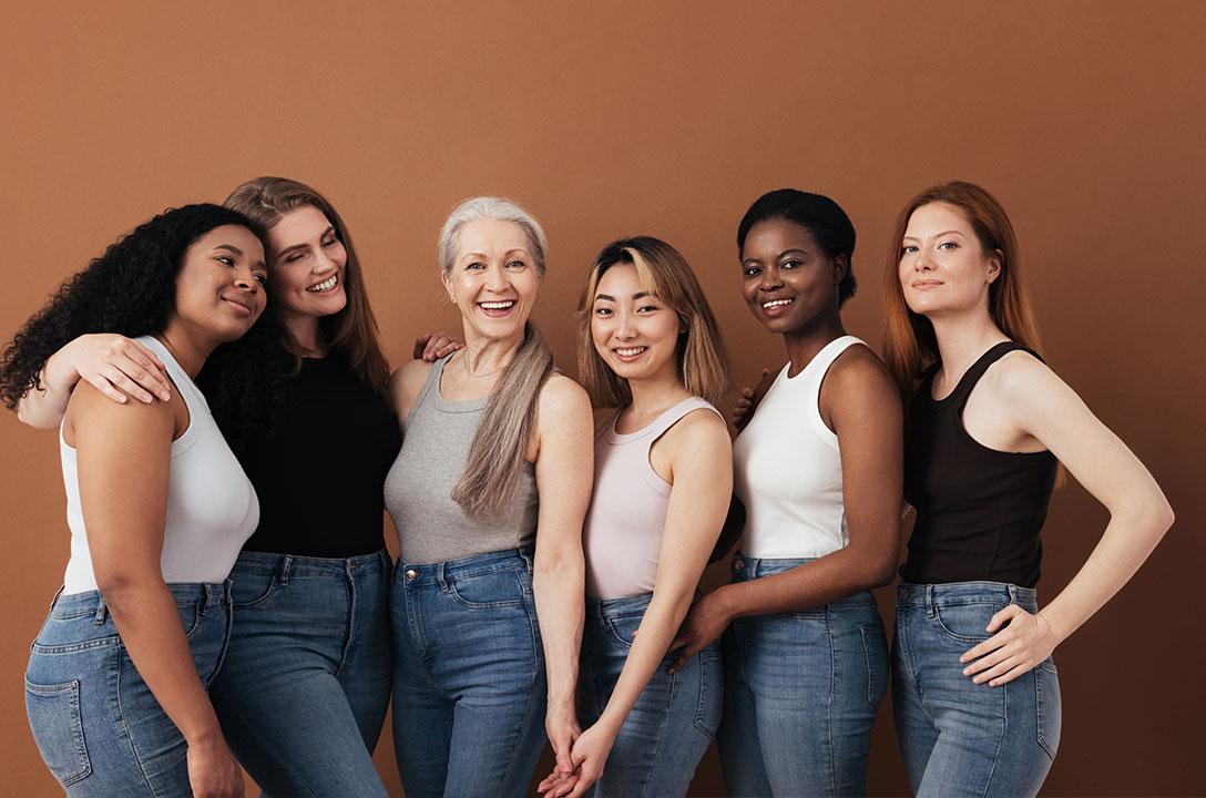 Six women of varied ages and skin tones in tank tops and jeans stand smiling together.
