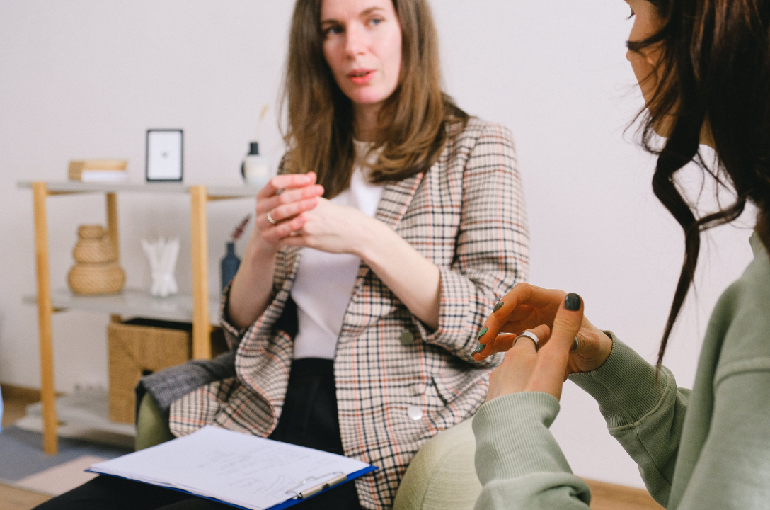Two women seated face to face in conversation, one in a plaid blazer, a clipboard of notes nearby.