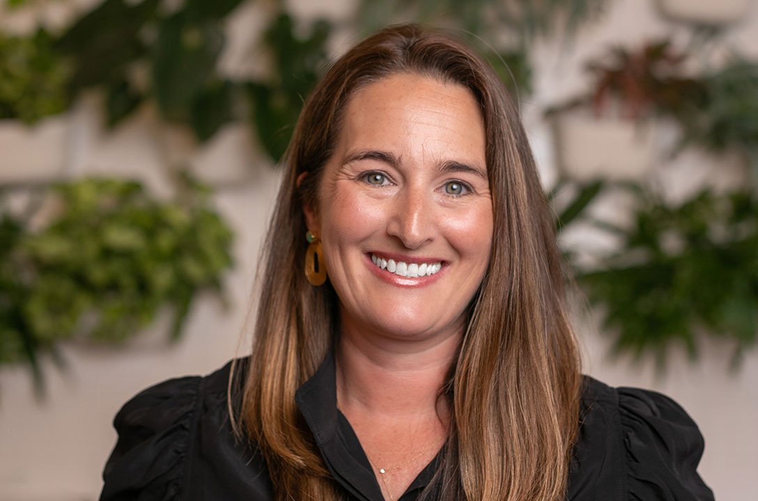 Smiling woman with long brown hair and gold earrings in a black blouse, leafy plants behind her.