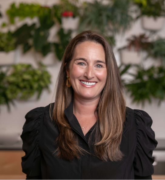 Smiling woman with long brown hair and gold earrings in a black blouse, leafy plants behind her.