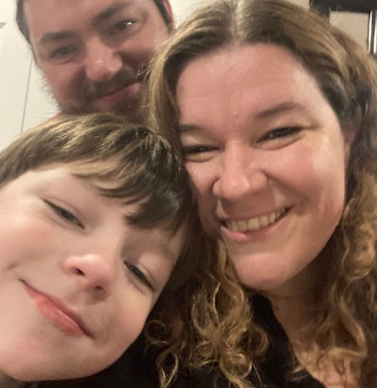 A smiling woman, a young child, and a bearded man pose together for a selfie outdoors.