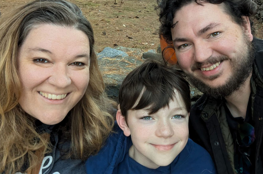 A smiling woman, a young child, and a bearded man pose together for a selfie outdoors.