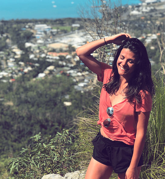 A smiling woman in a coral top stands on a grassy hilltop above a coastal town and sea.