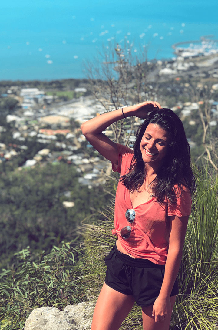 A smiling woman in a coral top stands on a grassy hilltop above a coastal town and sea.