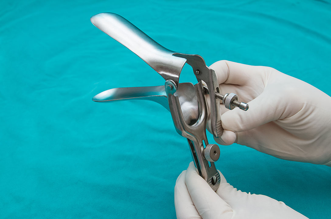 Gloved hands hold a stainless steel vaginal speculum over a teal cloth.