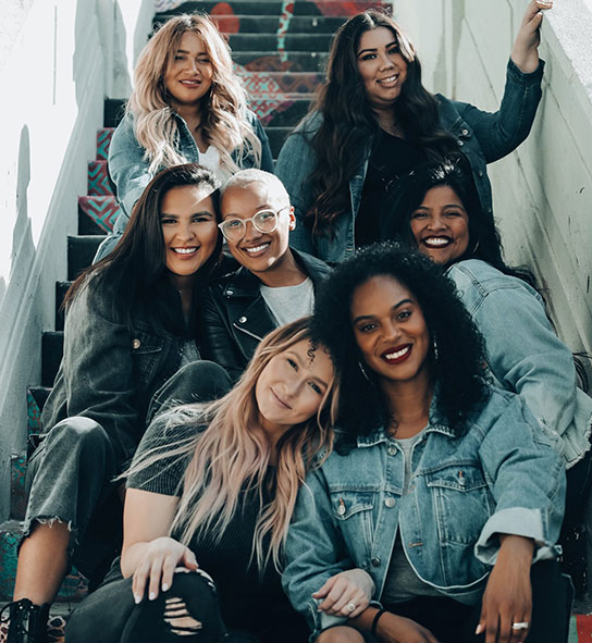 Seven smiling women in denim pose together on colorful painted outdoor stairs.