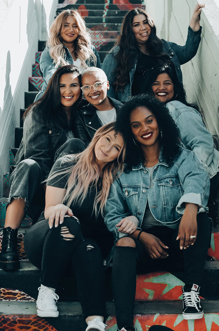 Seven smiling women in denim pose together on colorful painted outdoor stairs.