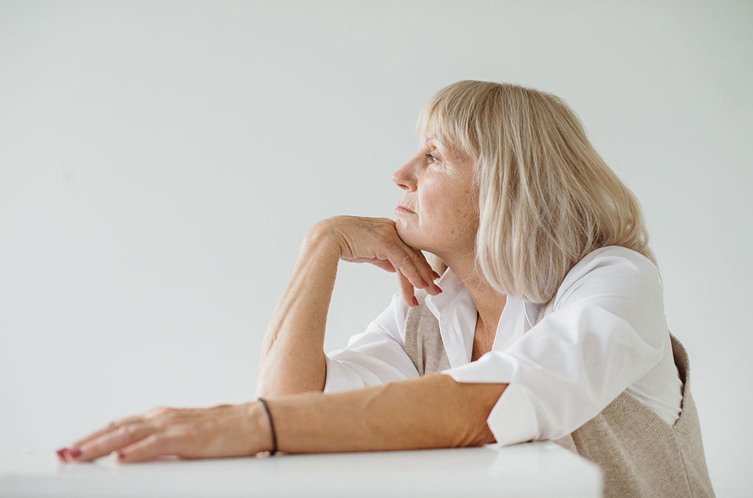 An older woman with light blonde hair rests her chin on her hand, gazing thoughtfully to the side.