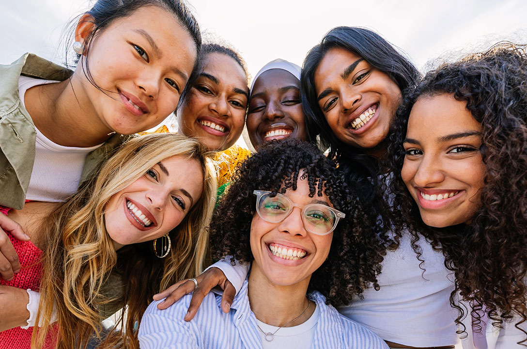 A close group of seven smiling young women of diverse backgrounds huddled together outdoors.
