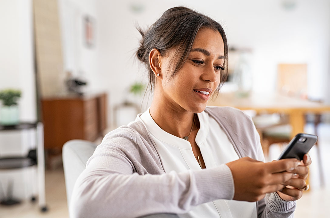 A woman in a cardigan sits indoors, smiling while looking at her smartphone.
