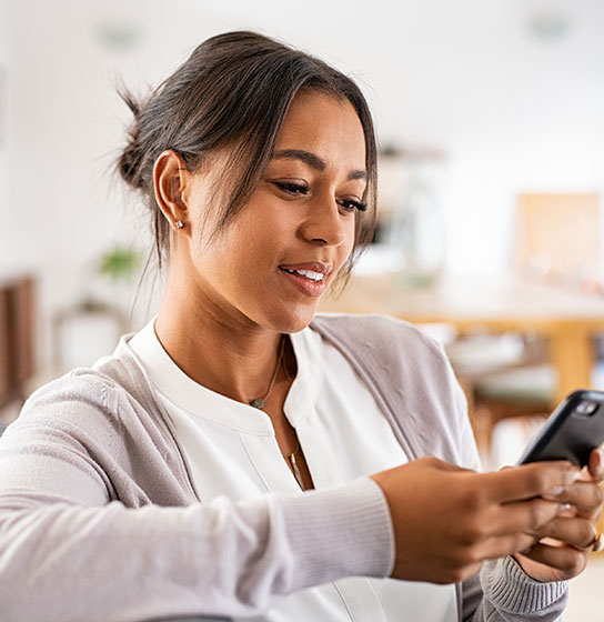 A woman in a cardigan sits indoors, smiling while looking at her smartphone.