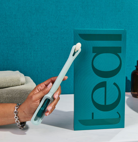 A hand holds the teal wand device beside its 'teal' box on a bathroom counter with flowers and a sink.