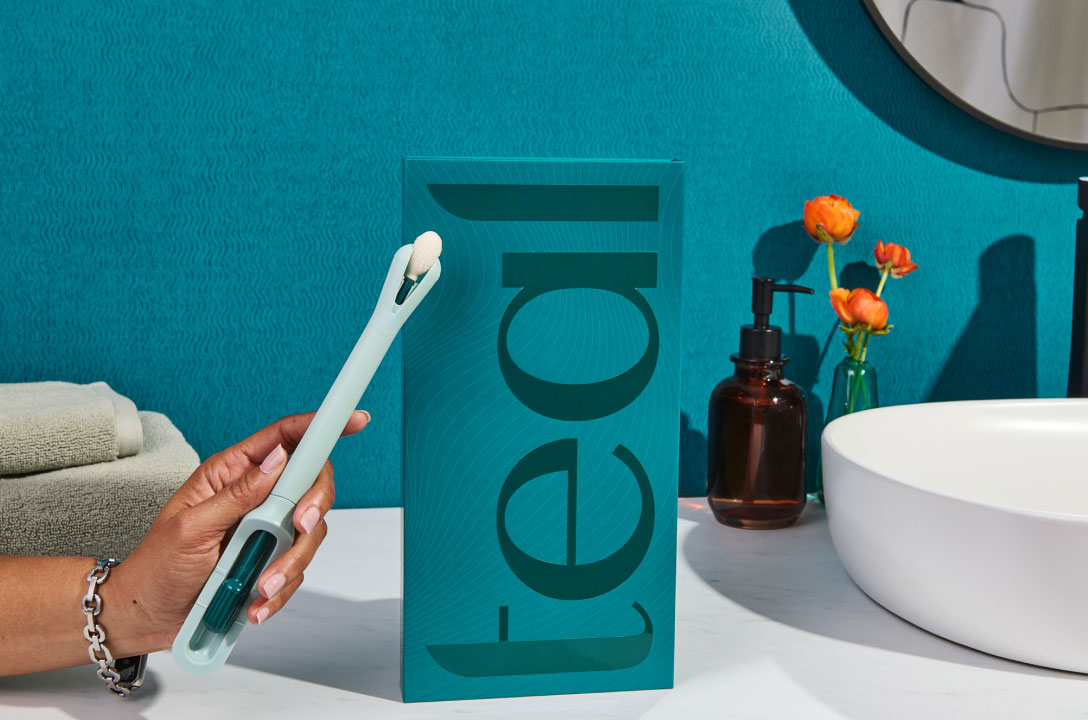 A hand holds the teal wand device beside its 'teal' box on a bathroom counter with flowers and a sink.
