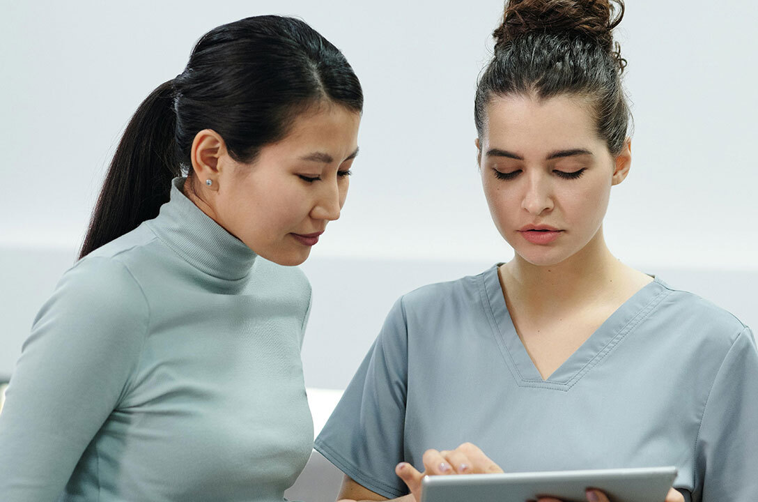 Two women in gray tops, one in medical scrubs, look down together at a tablet one is holding.