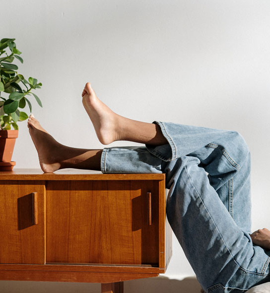 Bare feet and jeans-clad legs propped on a wooden credenza beside a potted plant.