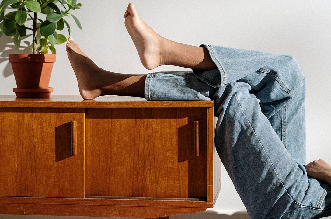Bare feet and jeans-clad legs propped on a wooden credenza beside a potted plant.