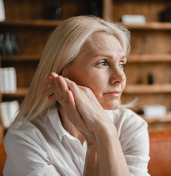 Blonde woman in a white shirt rests her chin on her hands, gazing away, bookshelves behind her.