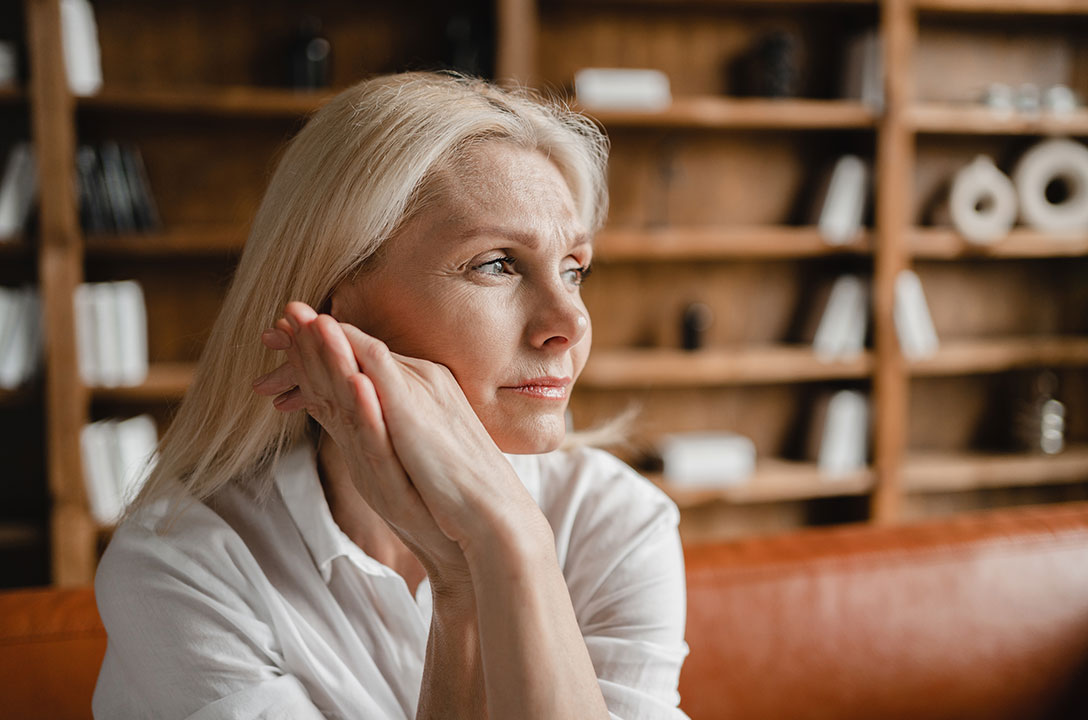 Blonde woman in a white shirt rests her chin on her hands, gazing away, bookshelves behind her.