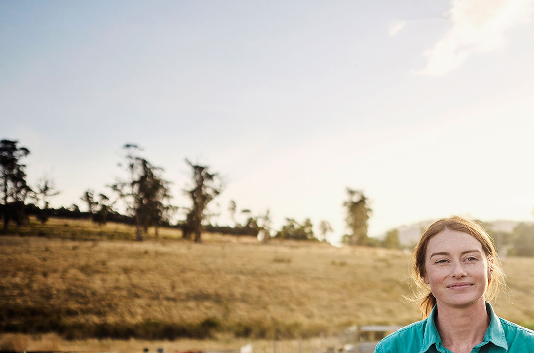 Woman in a teal shirt smiles outdoors before a dry grassy hillside dotted with trees at sunset.