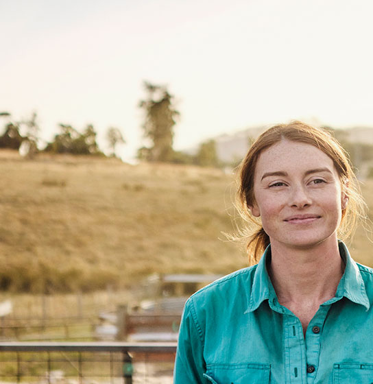 Woman in a teal shirt smiles outdoors before a dry grassy hillside dotted with trees at sunset.