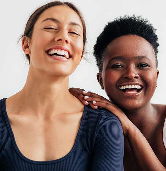 Two women smile against a white wall, one resting a hand on the other's shoulder.