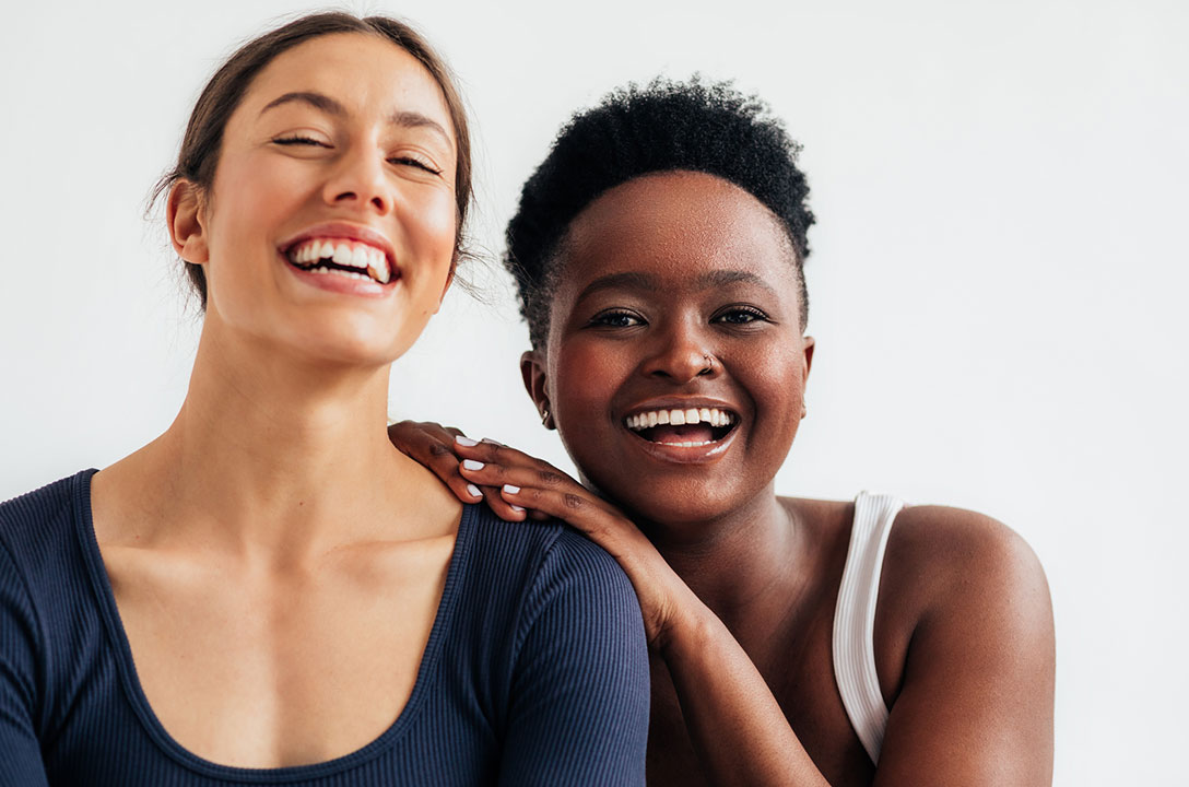 Two women smile against a white wall, one resting a hand on the other's shoulder.