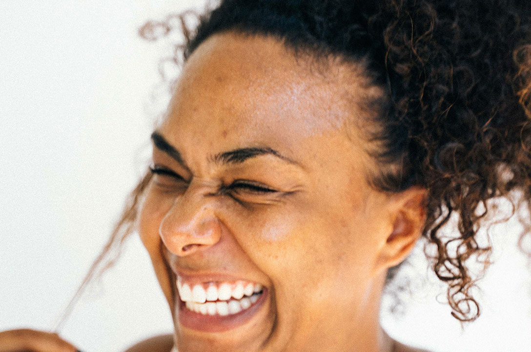 Close-up of a woman with curly hair laughing with her eyes closed against a white background.