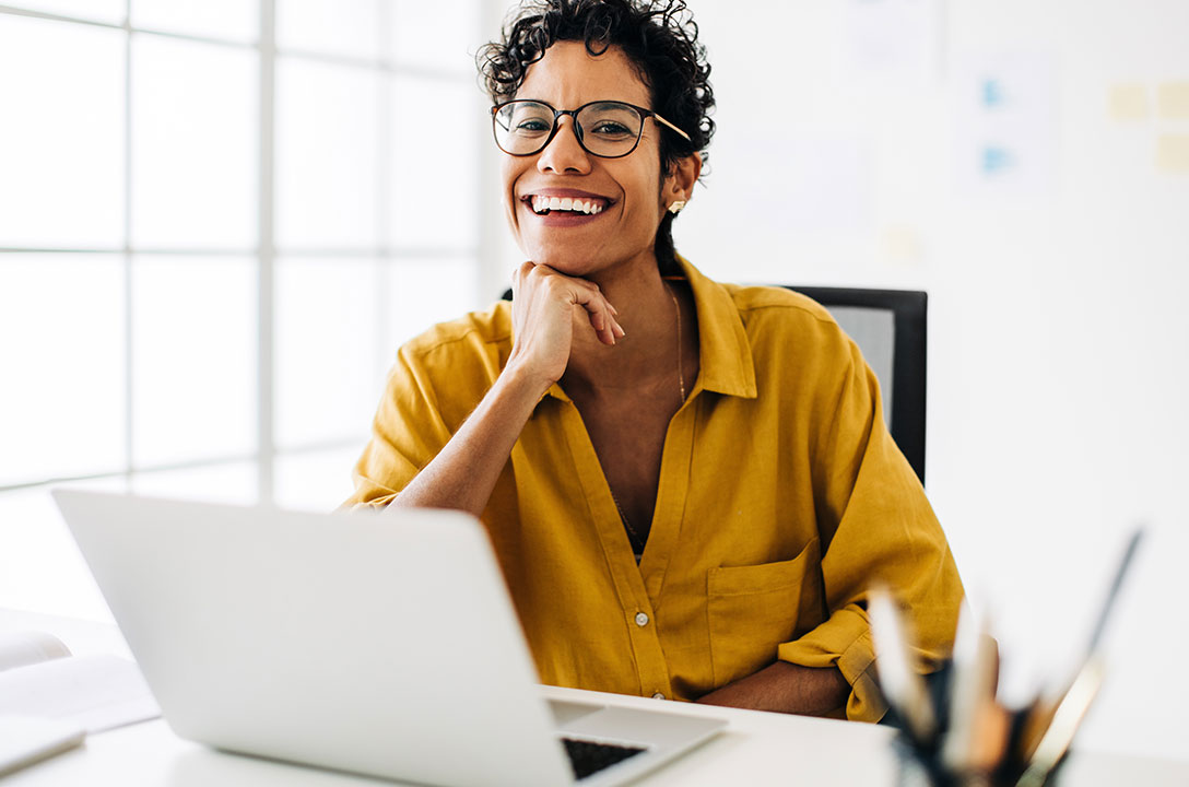 Woman in glasses and a yellow shirt smiles with her chin on her hand at a desk with a laptop.